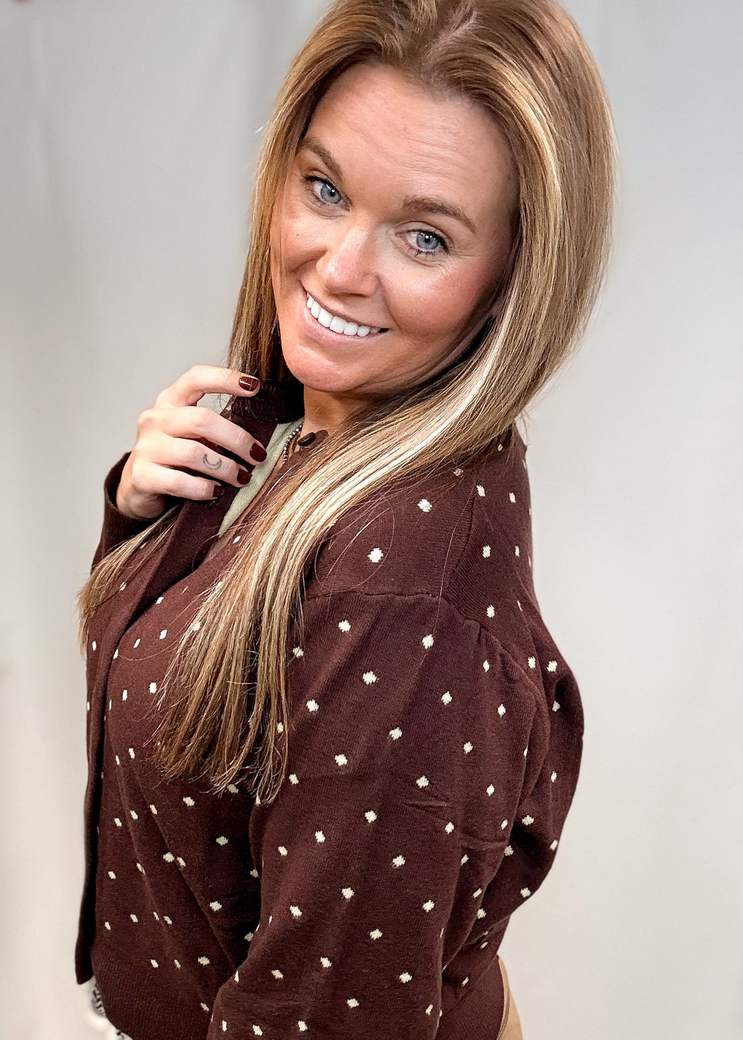 A smiling woman with blonde hair wearing a brown long-sleeve top with small white polka dots, photographed against a plain light gray background.