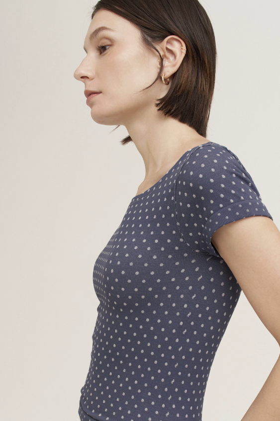 A woman shown in profile wearing a short-sleeve top with a navy blue base and small white polka dots, against a plain light beige studio background.