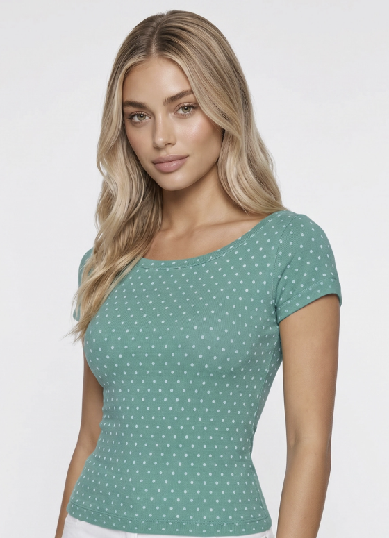 A woman wearing a short-sleeve scoop-neck top in sage green with a small white polka dot pattern, photographed against a light gray studio background.