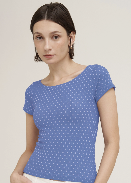 A woman wearing a blue short-sleeve top with a white polka dot pattern, photographed against a plain light beige studio background.
