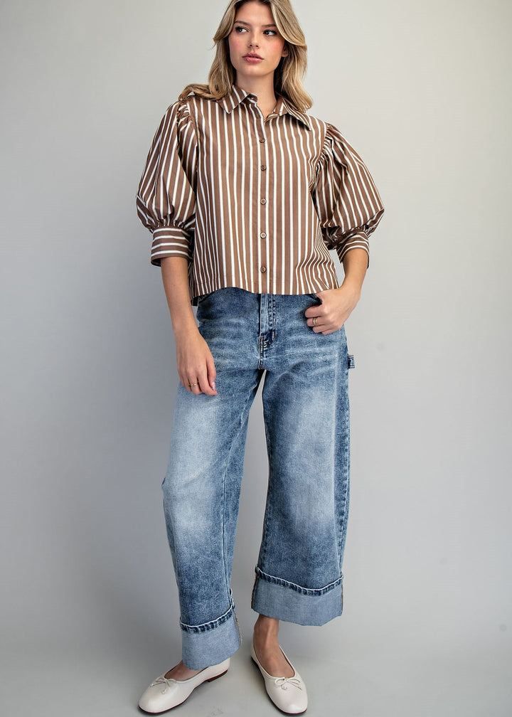 A woman stands against a light gray studio background wearing a brown and beige vertically striped button-up shirt with voluminous sleeves, blue wide-leg cropped jeans, and white flats.