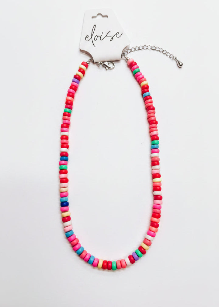 A colorful beaded necklace made of small cylindrical beads in pink, red, orange, yellow, green, blue, and white, displayed on a white surface with a branded card that reads &