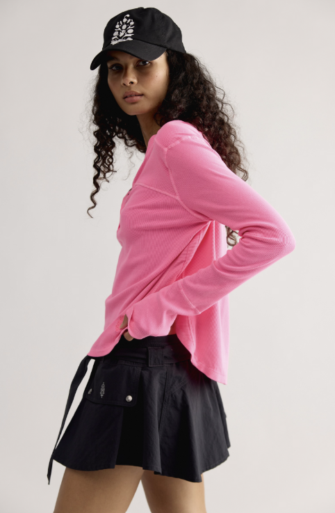 A model wearing a bright pink ribbed long-sleeve crop top, a black cap with a white logo, and a black pleated mini skirt, posed against a light gray studio background.