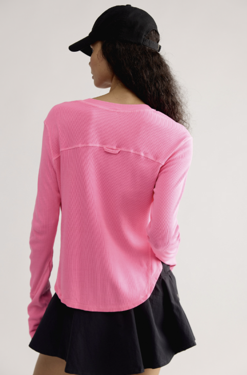 A woman shown from the back wearing a bright pink long-sleeve textured top, a black cap, and a black skirt, against a plain light gray studio background.