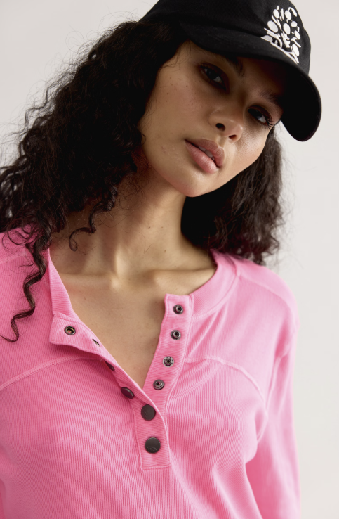 A close-up of a woman wearing a bright pink ribbed Henley shirt with dark buttons and a black baseball cap with a white graphic logo.