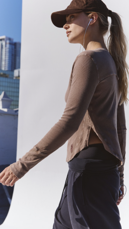 A woman in a side profile wearing a brown textured long-sleeve crop top, a brown baseball cap, and dark gray athletic pants, with white wired earphones in her ear.