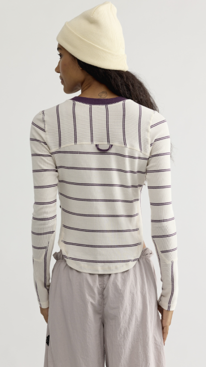 A model shown from the back wearing a cream ribbed long-sleeve top with thin dark purple horizontal stripes, a matching cream knit beanie, and light gray pants against a light gray studio background.