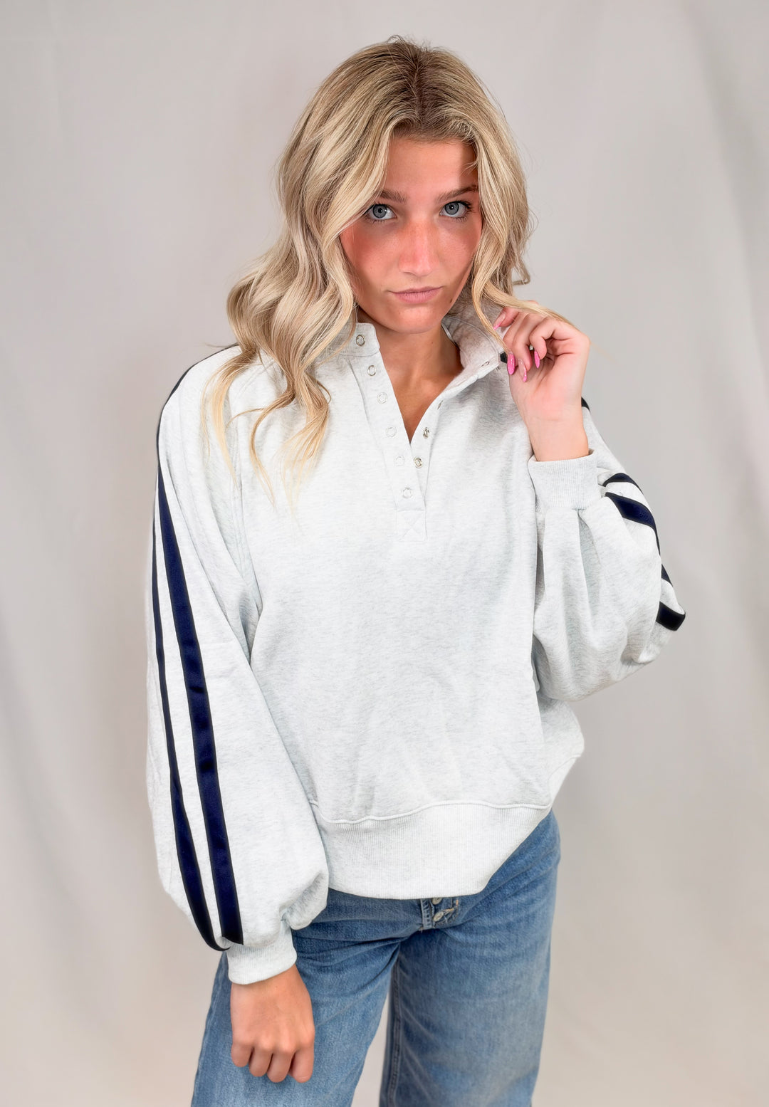 Woman wearing grey sweatshirt with navy stripes down sleeves with jeans on plain background