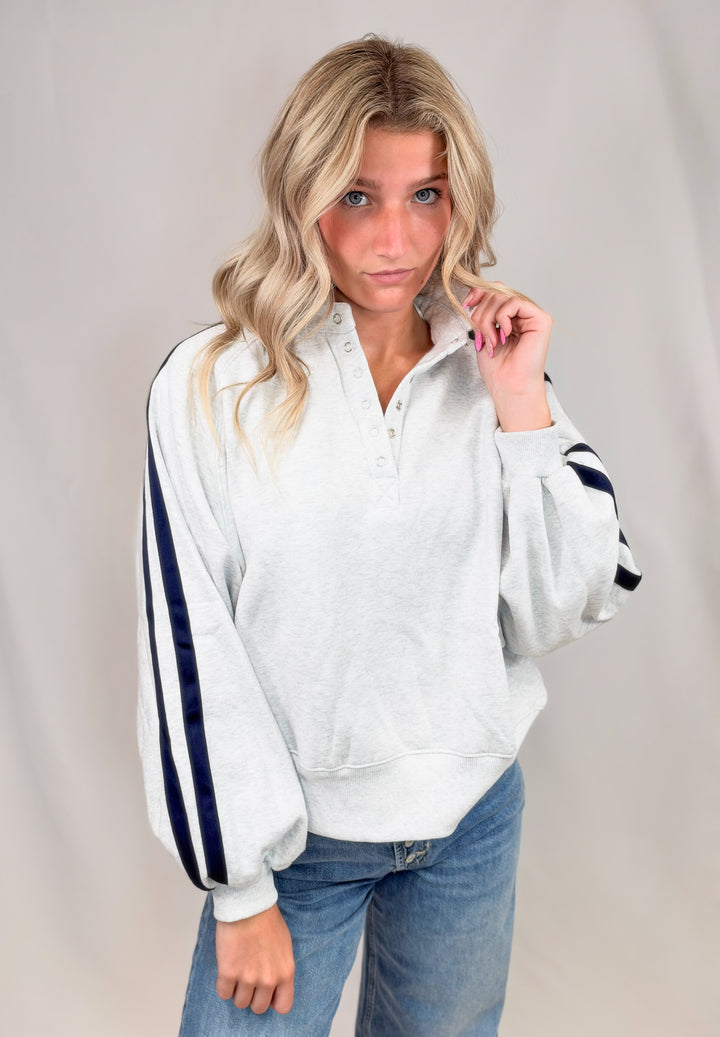 Woman wearing grey sweatshirt with navy stripes down sleeves with jeans on plain background