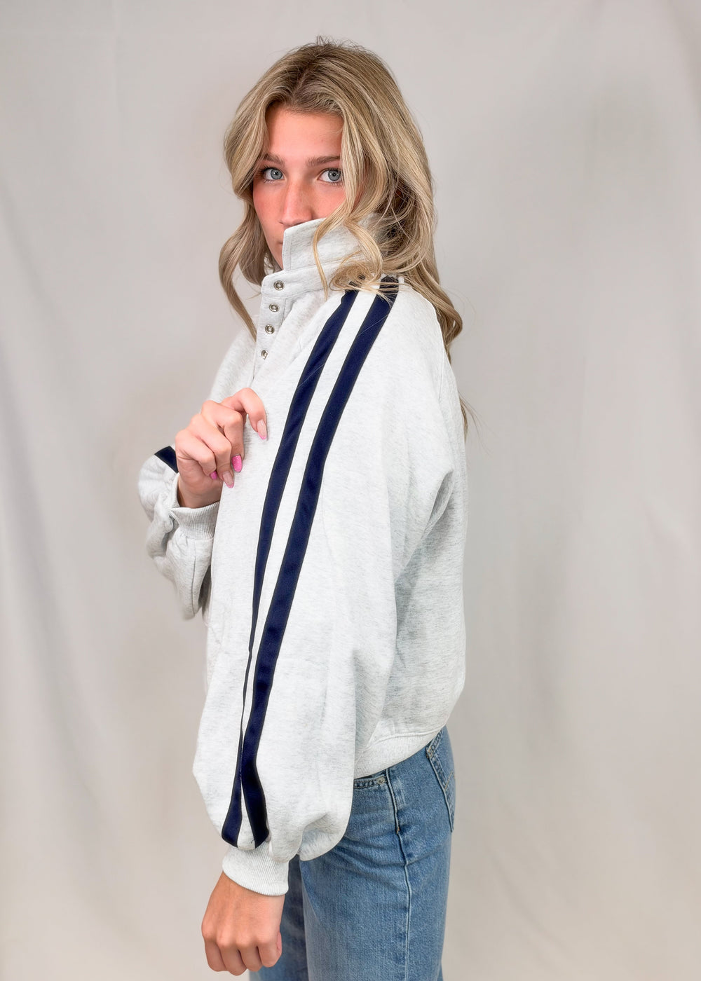 Woman shown from side view wearing grey sweatshirt with navy stripes down sleeves and jeans on plain background