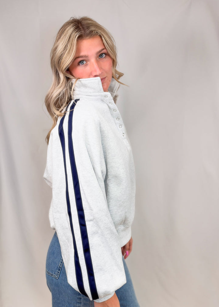 Woman shown from side view wearing grey sweatshirt with navy stripes down sleeves and jeans on plain background