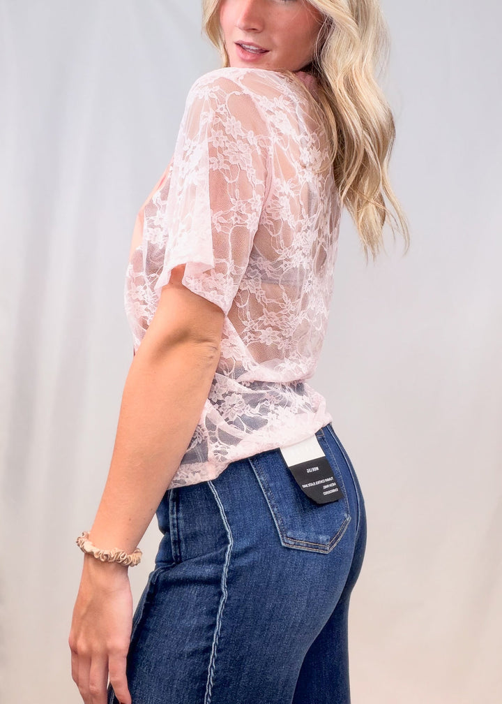 Woman shown from side view wearing pink lace tee with denim jeans on plain background