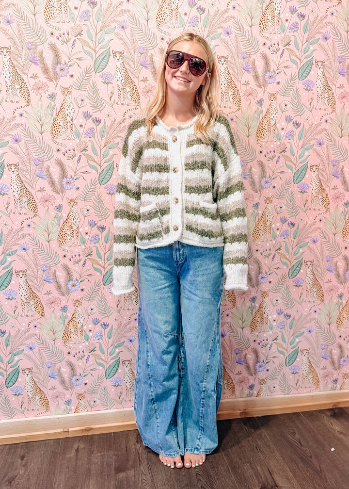A woman wearing a green and white striped fuzzy knit cardigan, blue wide-leg jeans, and sunglasses stands barefoot in front of pink patterned wallpaper with a wood floor.