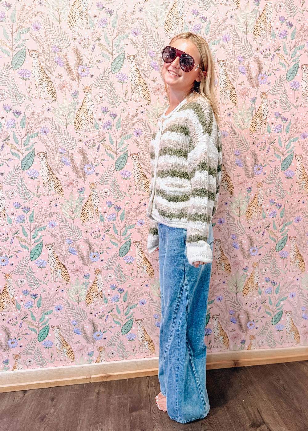 A woman wearing a fuzzy green and white striped cardigan, blue wide-leg jeans, and sunglasses stands barefoot in front of a pink patterned wallpaper wall.