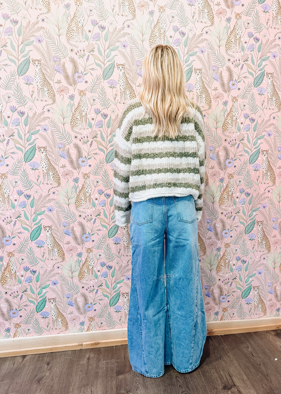 A woman stands with her back to the camera wearing a green and white striped knit sweater and wide-leg blue denim jeans, set against a pink patterned wallpaper background.
