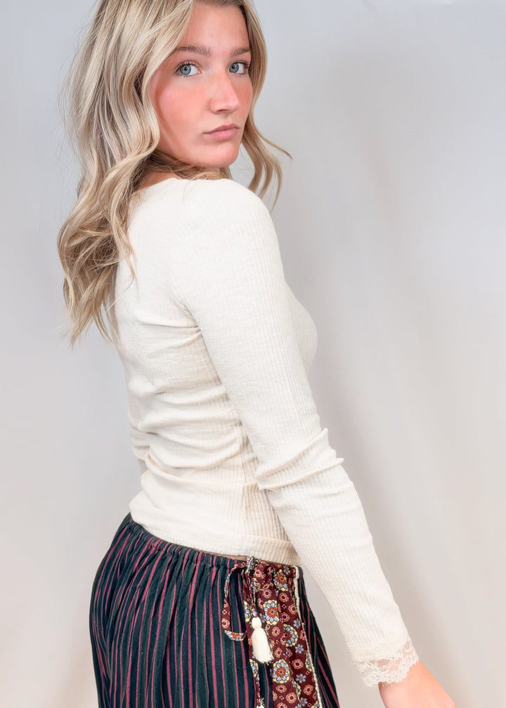 Woman shown from side view wearing cream ribbed long sleeve top with lace detail on neck and cuffs on plain background