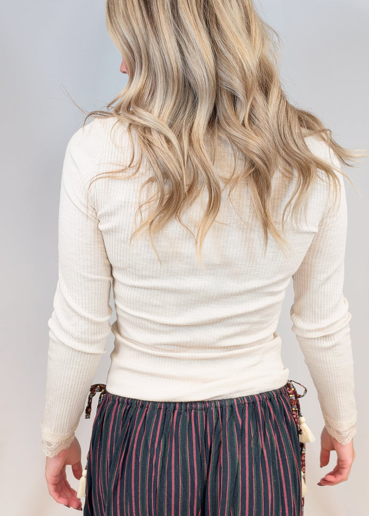 Woman shown from back view wearing cream ribbed long sleeve top with lace detail on cuffs on plain background