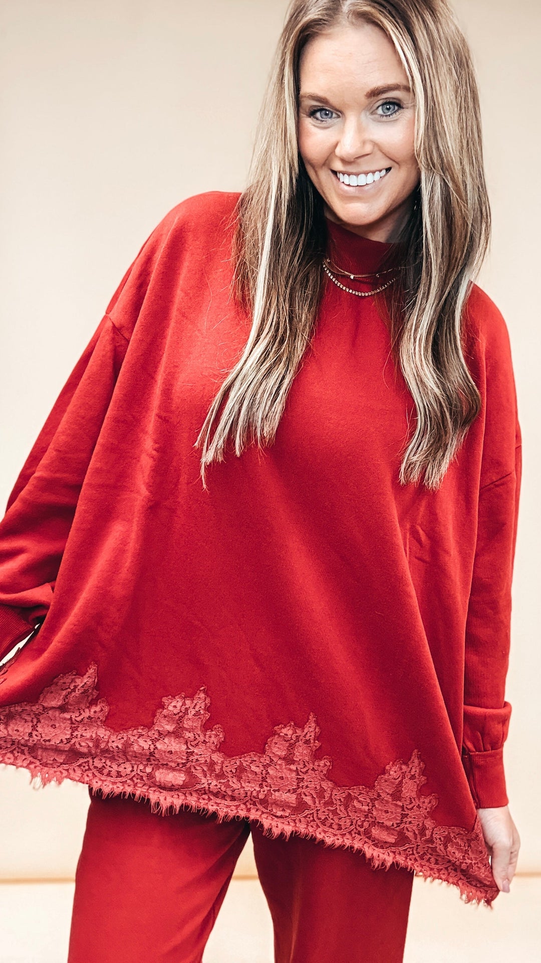 A smiling woman wearing a burgundy long-sleeve knit poncho-style top with matching red pants, photographed against a plain light beige studio background.