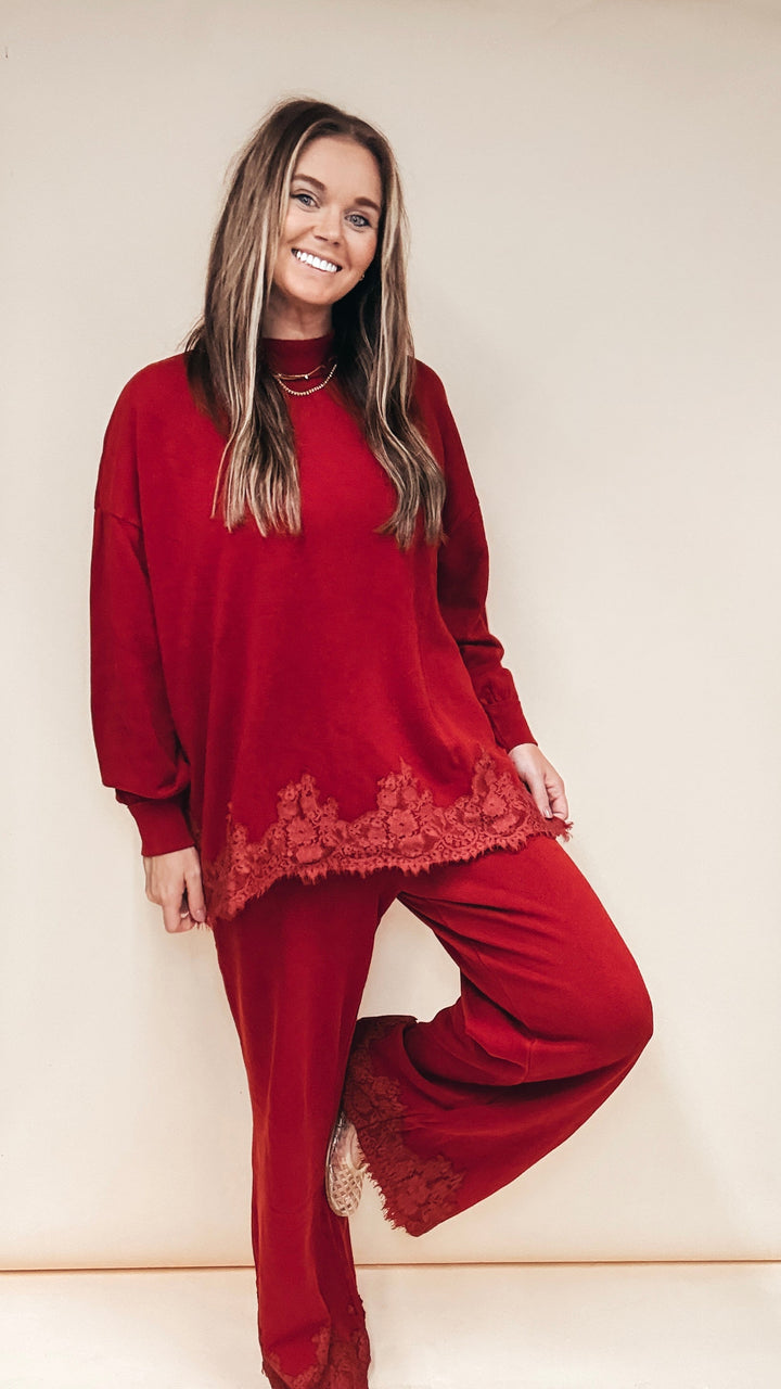 A woman modeling a matching red lounge set consisting of a long-sleeve top with lace trim and wide-leg pants, posed against a plain beige studio background.
