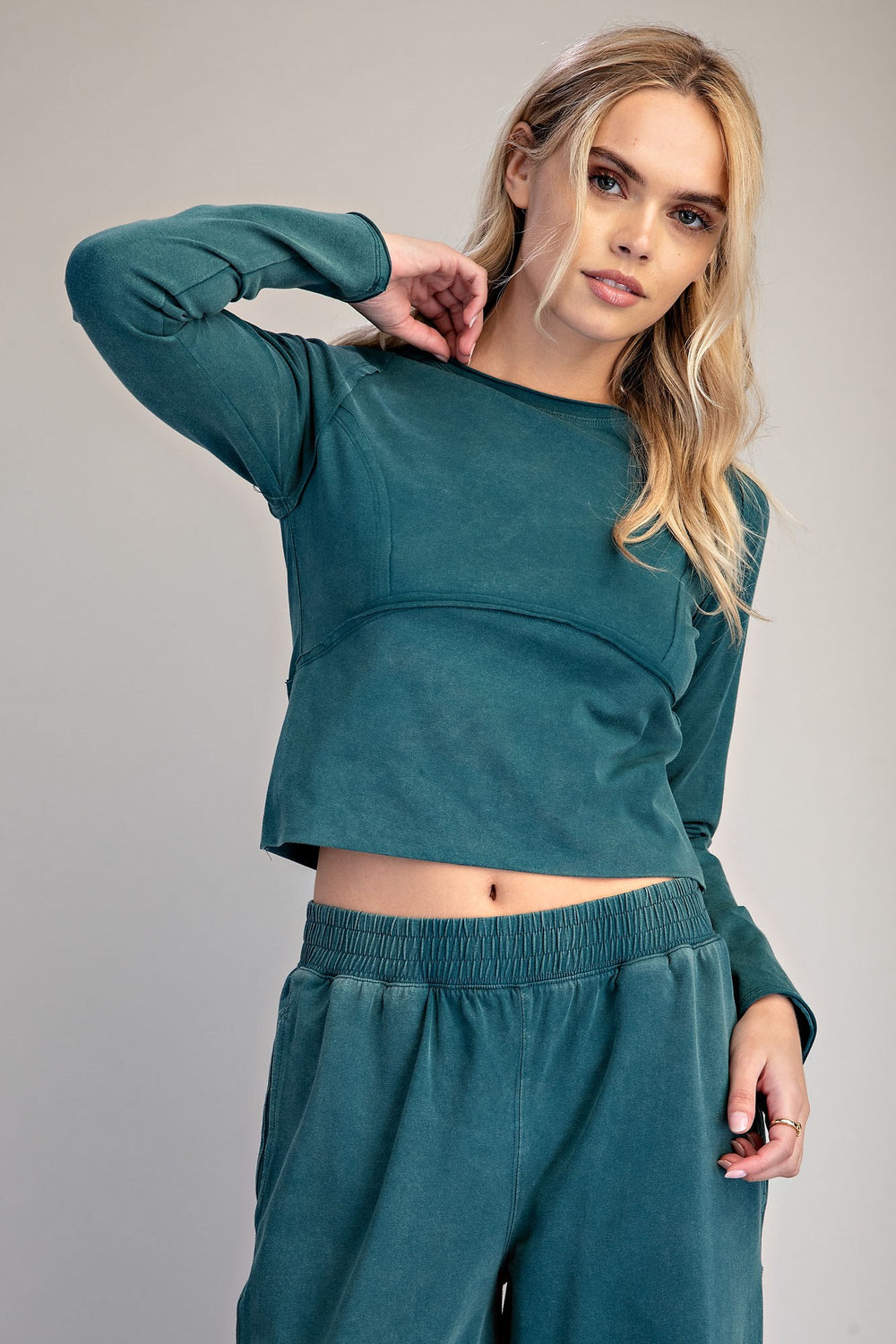 A woman modeling a matching two-piece lounge set in a dusty teal color, featuring a long-sleeve cropped top and matching wide-leg sweatpants, against a solid light gray background.