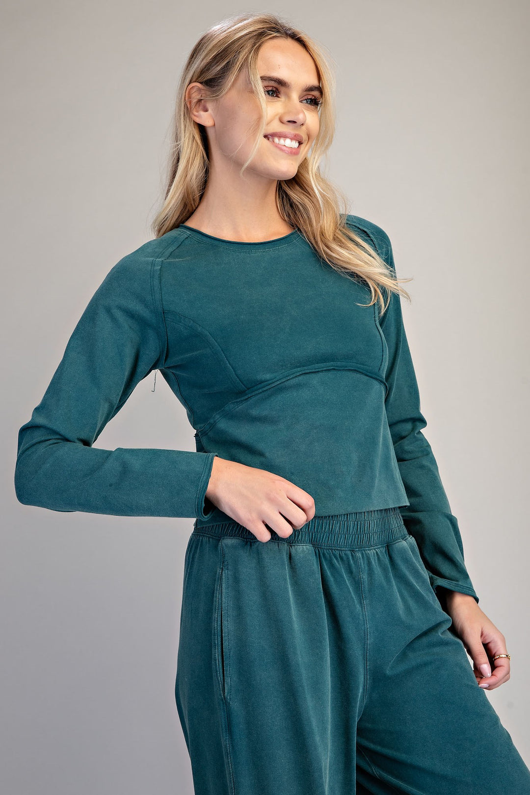 A woman modeling a matching two-piece lounge set in a deep teal color, featuring a long-sleeve crewneck top and matching wide-leg pants, set against a plain gray studio background.