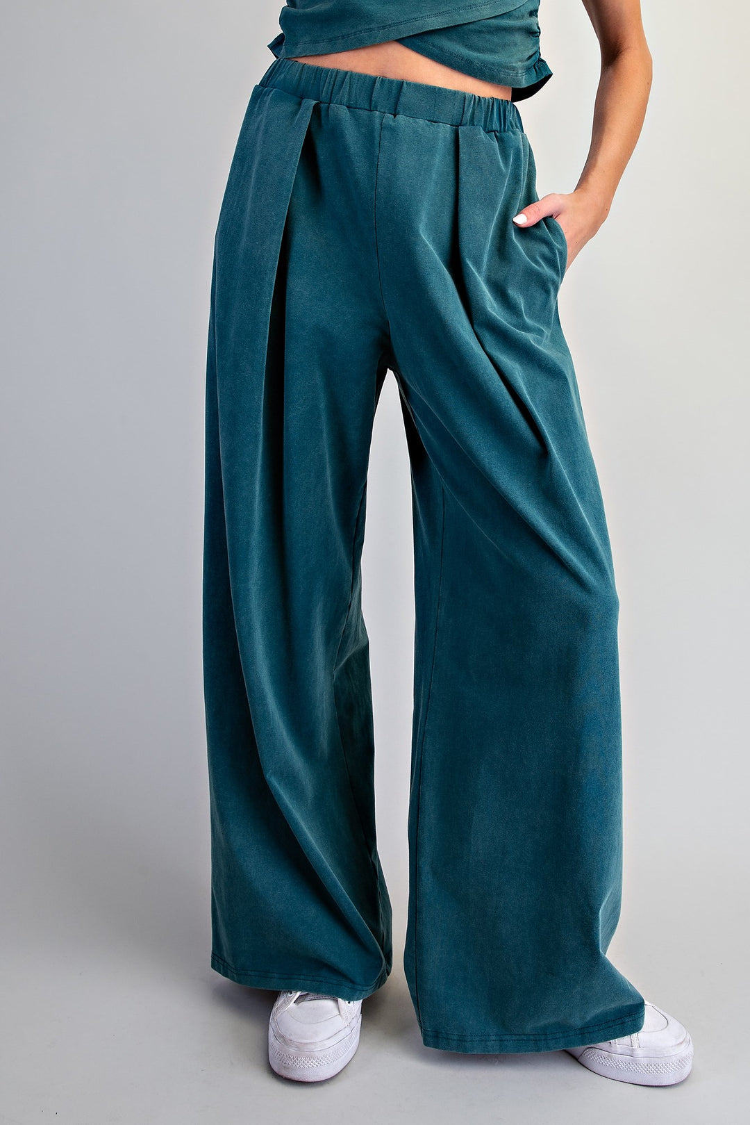 A person wearing high-waisted, wide-leg teal corduroy pants with an elastic waistband, styled with white sneakers against a neutral gray studio background.