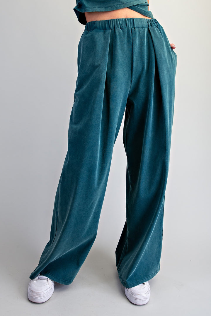 A person wearing wide-leg pleated trousers in a deep teal color, paired with white sneakers, shown from the waist down against a light gray studio background.