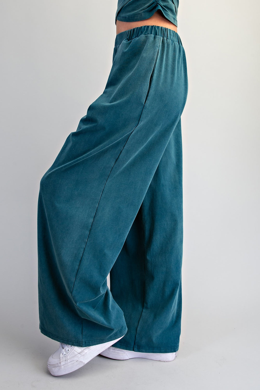 A person wearing wide-leg teal corduroy pants with an elastic waistband, paired with white sneakers, shown from the waist down against a light gray studio background.