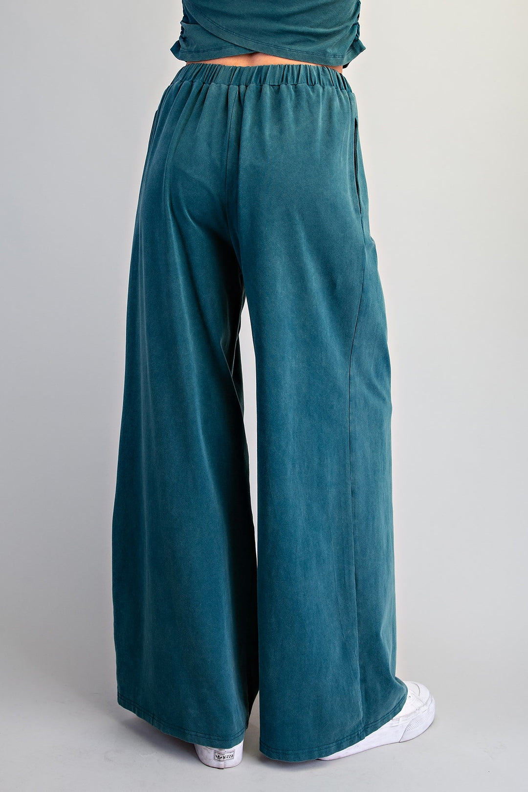 A person shown from the back wearing wide-leg teal pants and a matching teal top, standing against a light gray studio background.
