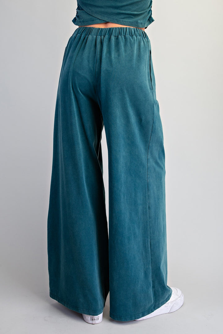 A person shown from the back wearing wide-leg teal pants and a matching teal top, standing against a light gray studio background.