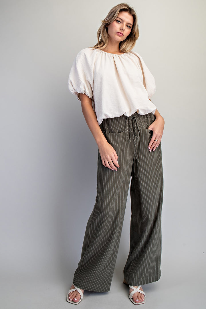 A woman modeling a white short-sleeve blouse with puff sleeves and dark olive green wide-leg pinstripe pants, paired with white heeled sandals, against a solid light gray studio background.