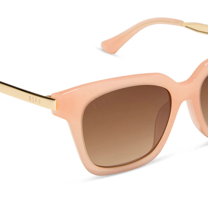 Close-up of a pair of sunglasses with a peach-pink frame, brown gradient lenses, and gold metal temples. The brand name &