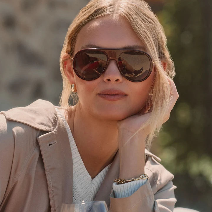 A close-up lifestyle shot of a blonde woman wearing dark brown aviator sunglasses with a gold-tone frame, a beige trench coat, and a white knit sweater.