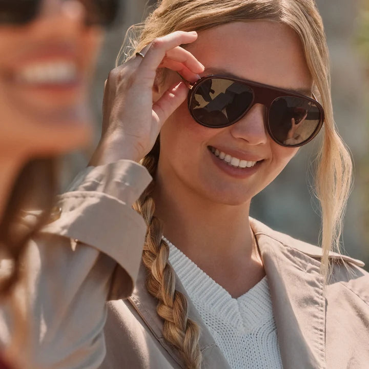A close-up lifestyle shot of a woman wearing dark brown aviator sunglasses with a beige trench coat and white knit sweater, adjusting the side of the sunglasses with her hand.