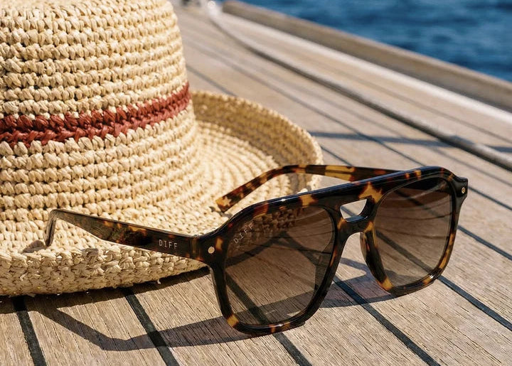 Pair of tortoise sunglasses with brown gradient lenses sitting near a hat outdoors