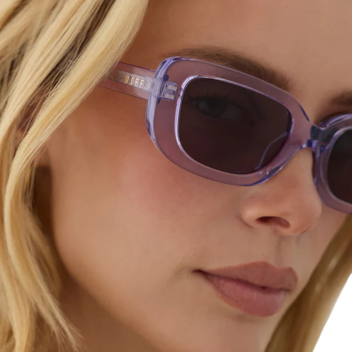Close-up of a model wearing translucent lavender-purple sunglasses with dark lenses and a visible DKNY logo on the temple arm.