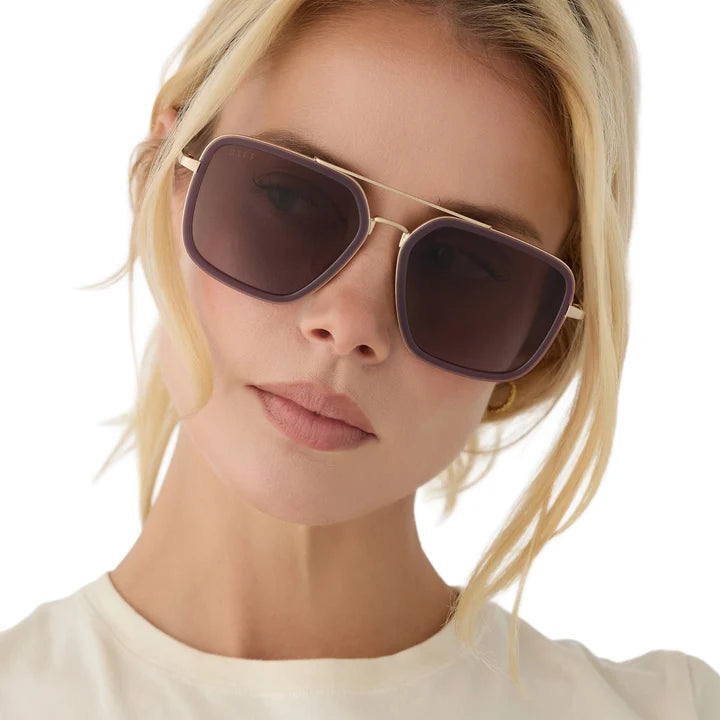 A close-up of a blonde woman wearing aviator-style sunglasses with a thin gold metal frame and dark brown lenses, set against a clean white background.