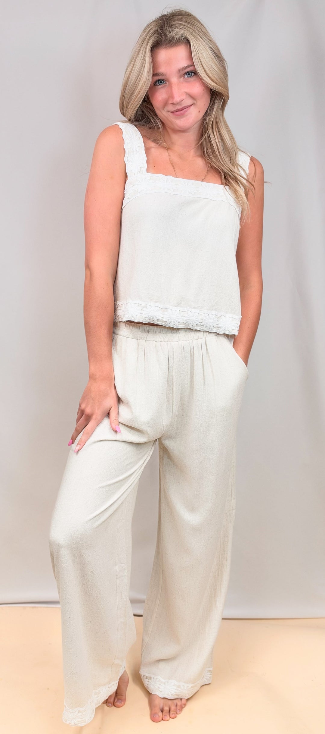 Woman wearing matching tank and pant linen set with floral details on sleeve, waist and ankle on plain background