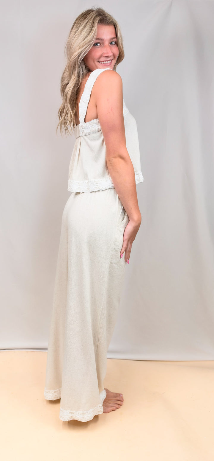 Woman shown from side view wearing matching tank and pant linen set with floral design on straps and ankles on plain background