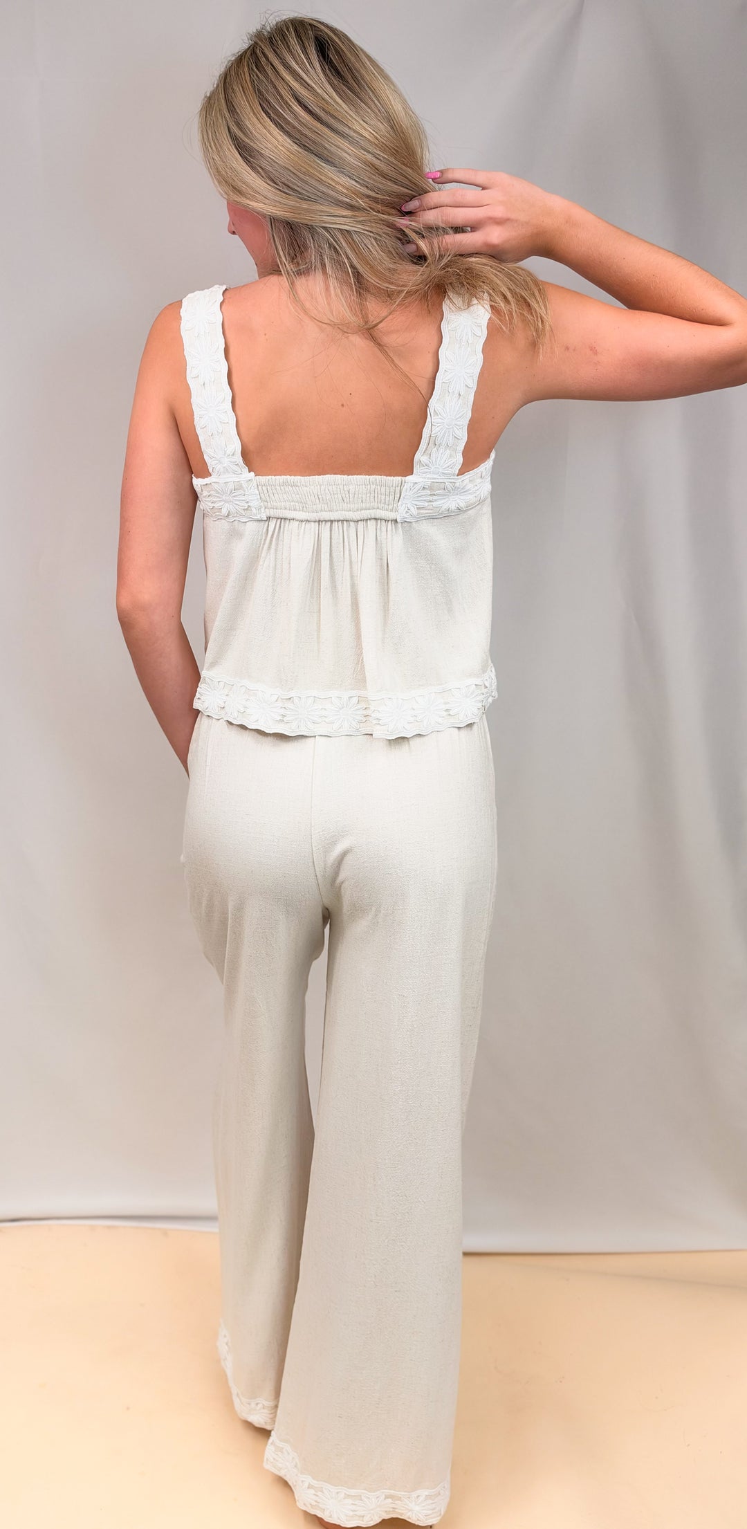 Woman shown from back view wearing matching tank and pant linen set with floral designs on straps and ankles on plain background