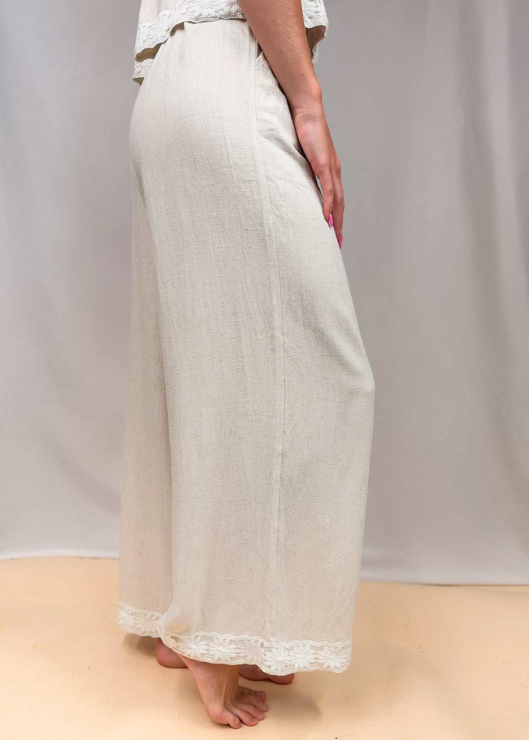 Woman shown waist down from side view wearing linen pants with floral detail on ankles on plain background