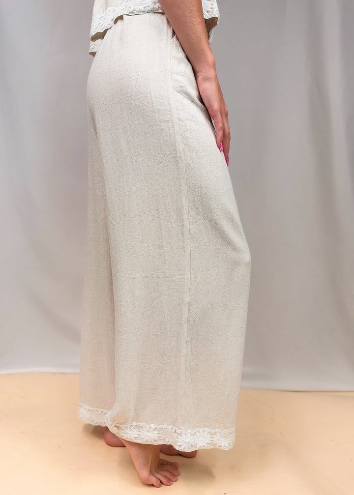 Woman shown waist down from side view wearing linen pants with floral detail on ankles on plain background