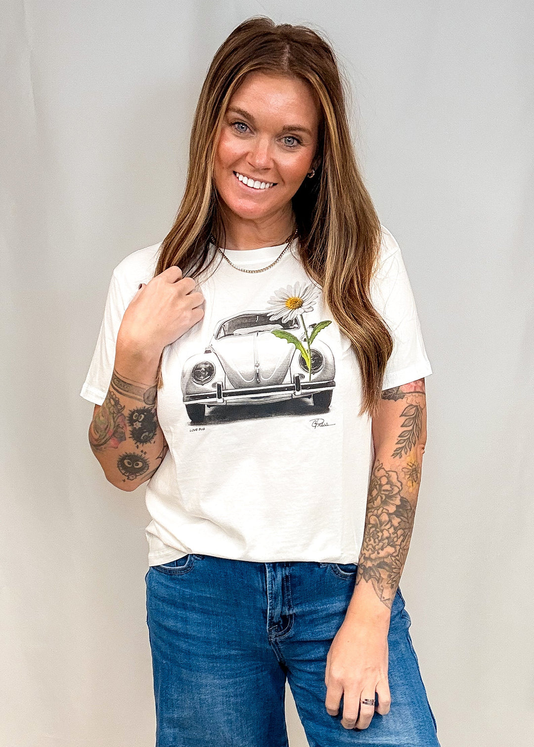 A woman with long brown hair and tattoos models a white graphic t-shirt featuring a vintage Volkswagen Beetle with a daisy, paired with blue jeans, against a light gray studio backdrop.