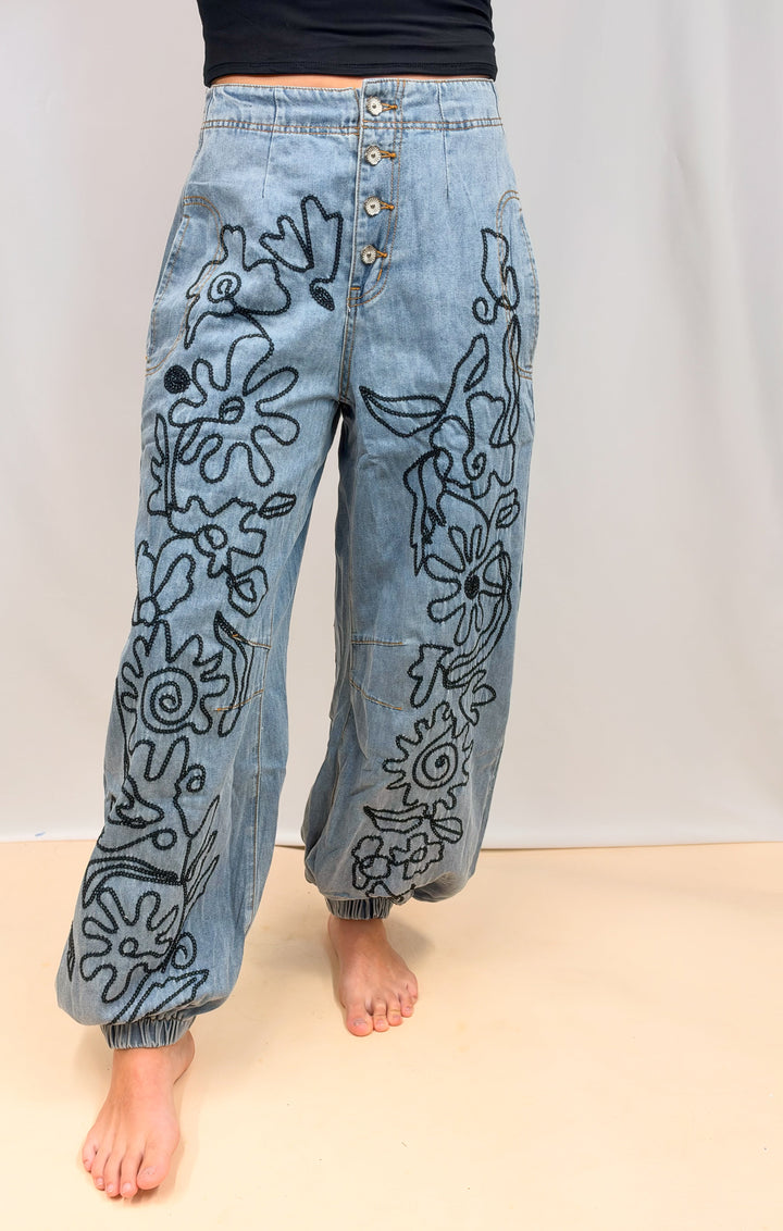 Light blue denim pants with black floral line-art embroidery, featuring a button-fly front and elastic cuffed harem-style legs, shown on a model against a neutral studio backdrop.
