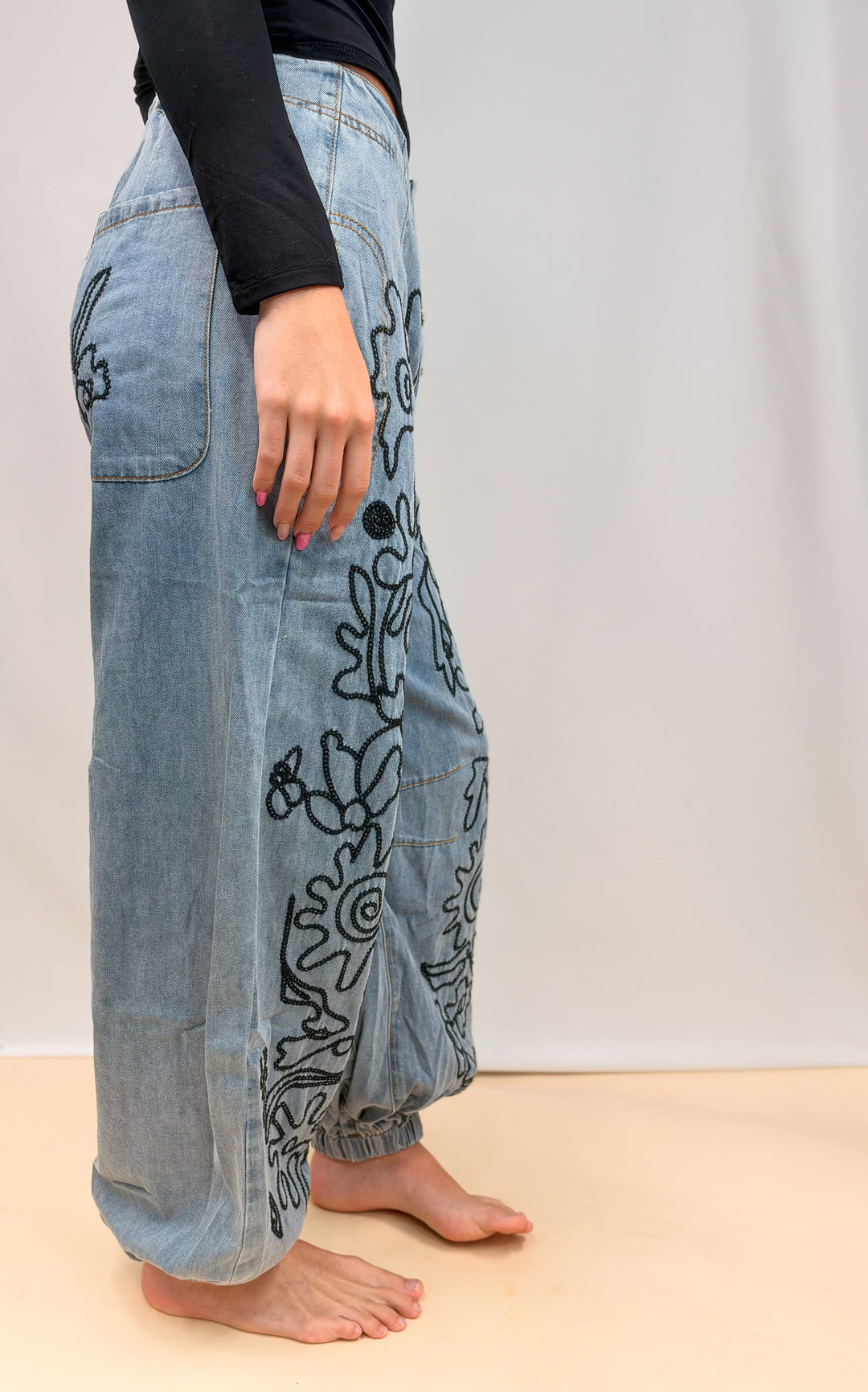 Woman shown from side view wearing denim pants with floral embroidery on plain background
