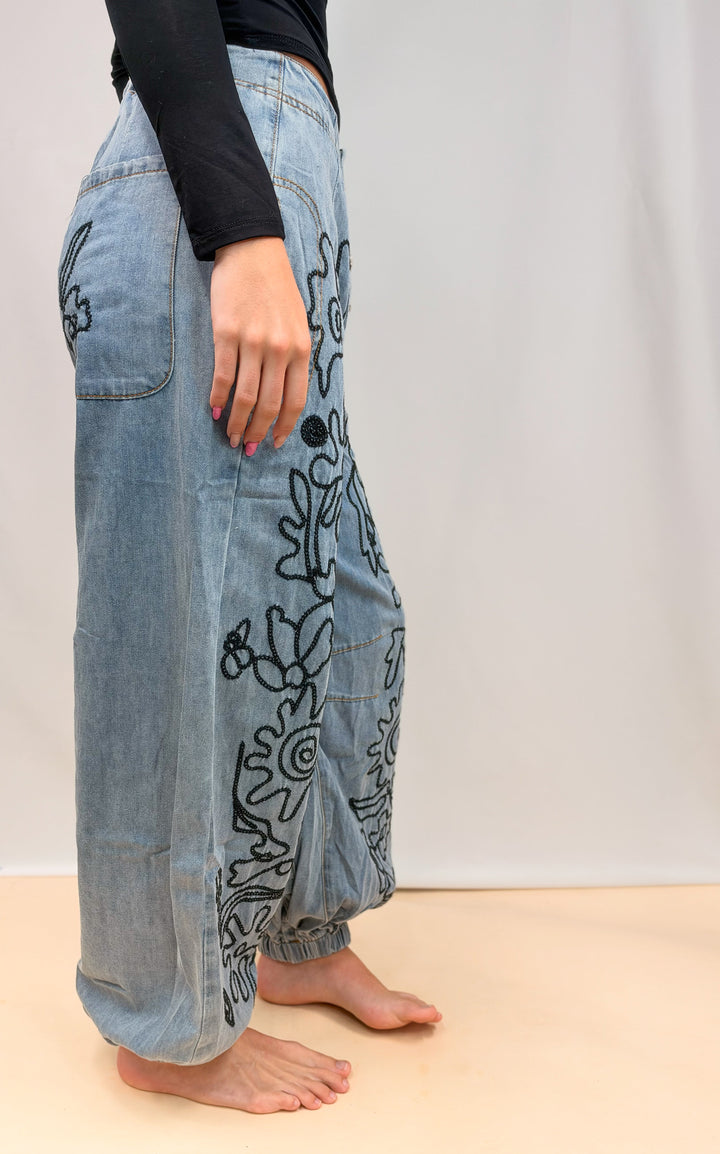 Woman shown from side view wearing denim pants with floral embroidery on plain background