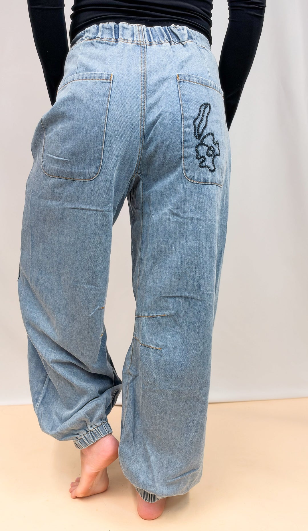 Woman shown from back view wearing denim joggers with floral embroidery on back pocket on plain background