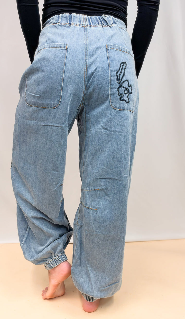 Woman shown from back view wearing denim joggers with floral embroidery on back pocket on plain background