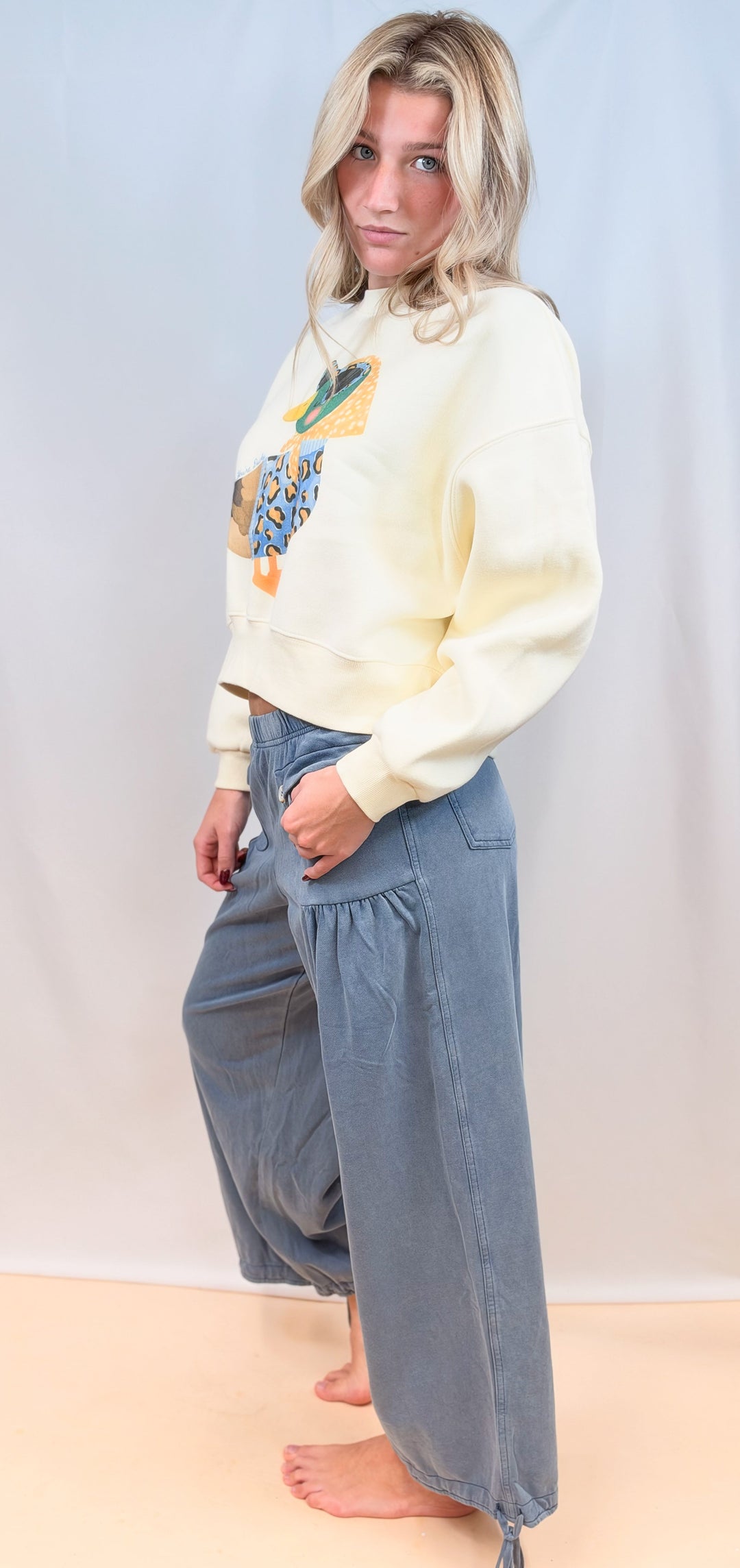 Woman shown from side view wearing cream cropped sweatshirt with duck graphic and blue pants on plain background