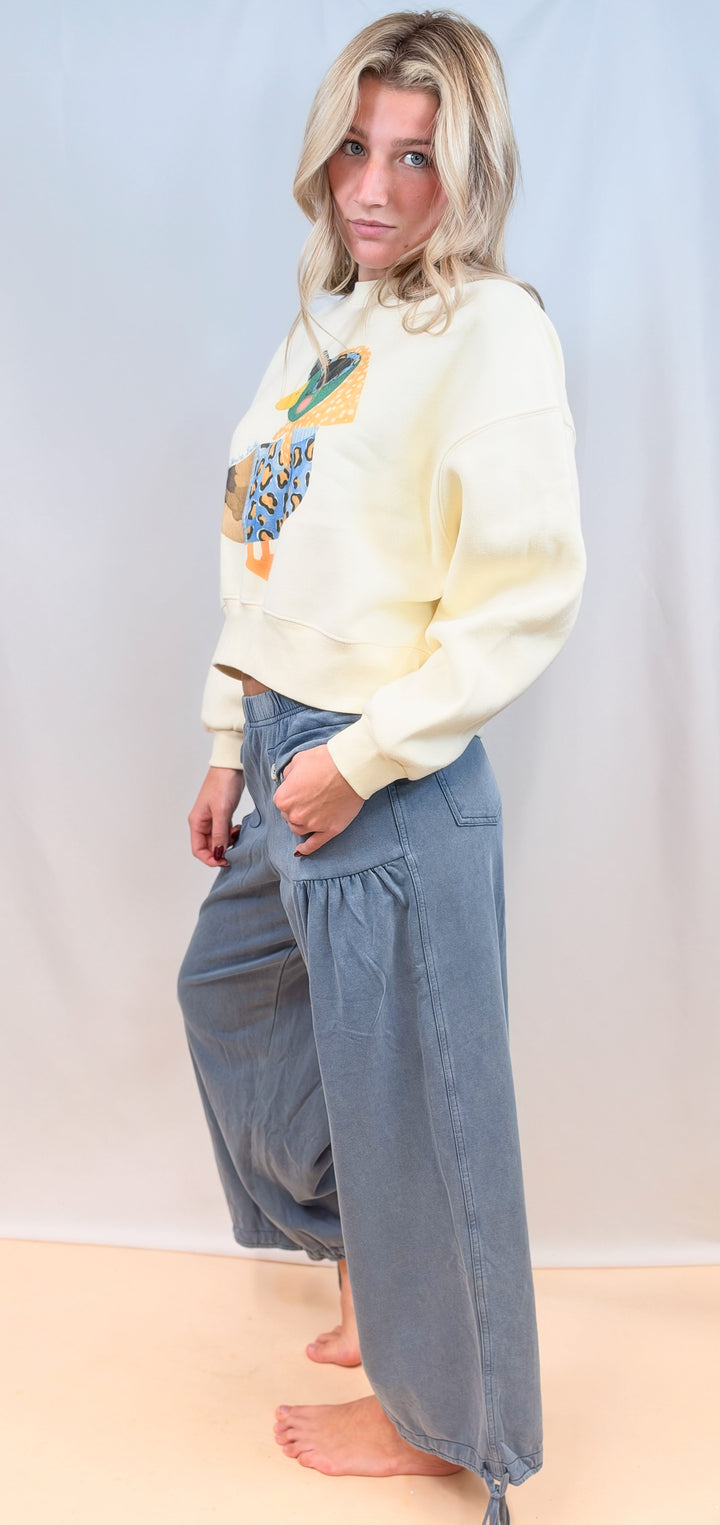 Woman shown from side view wearing cream cropped sweatshirt with duck graphic and blue pants on plain background